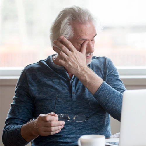 A man sitting in front of his computer rubbing his eye with one hand as the other holds his eyeglasses